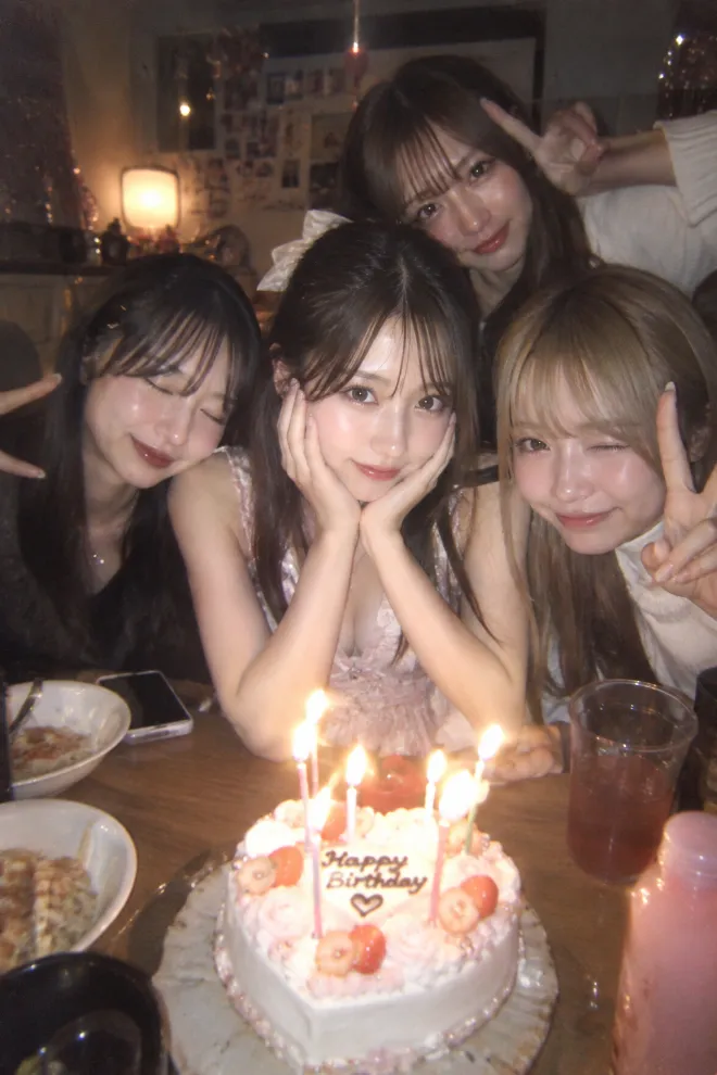 暗所のコンパクトデジタルカメラで撮ったような写真。フェミニンな服装の20歳の日本人女性が複数の友人と誕生日会を楽しんでいる。