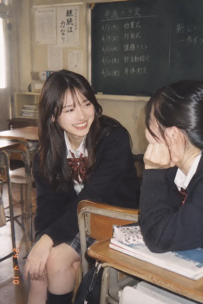 アナログ写真風。制服姿の20歳の日本人女性が教室で友人と会話している。