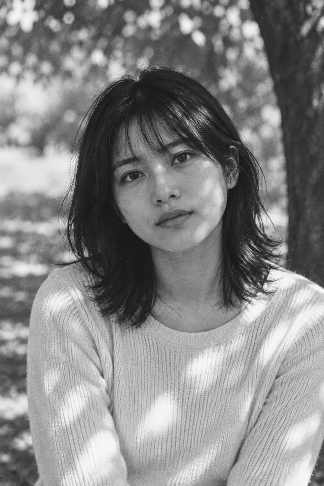 Kodak Tri-X 400で撮影したような白黒フィルム写真。シンプルな服装の20歳の日本人女性が木漏れ日の中でアンニュイな表情を見せている。