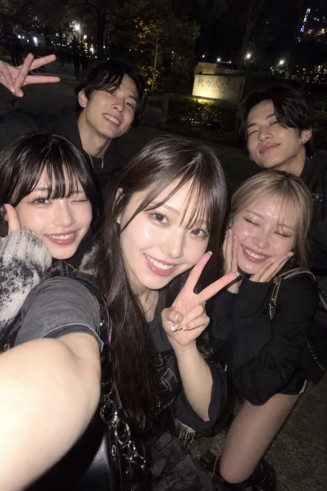 夜の直フラッシュ自撮り風。ストリート系ファッションの20歳の日本人女性が夜の公園で複数の友人と笑顔でセルフィーを撮っている。