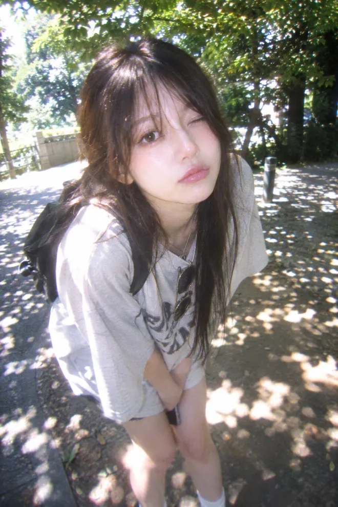 トイデジタルカメラで撮ったような写真。オーバーサイズの服を着た20歳の日本人女性が夏の木漏れ日の中にいる。