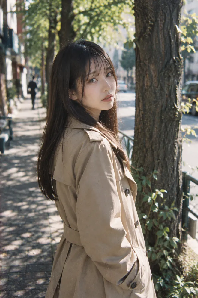 Leica M6で撮影したような35mmフィルム写真。トレンチコート姿の20歳の日本人女性が街路樹の前にいる。