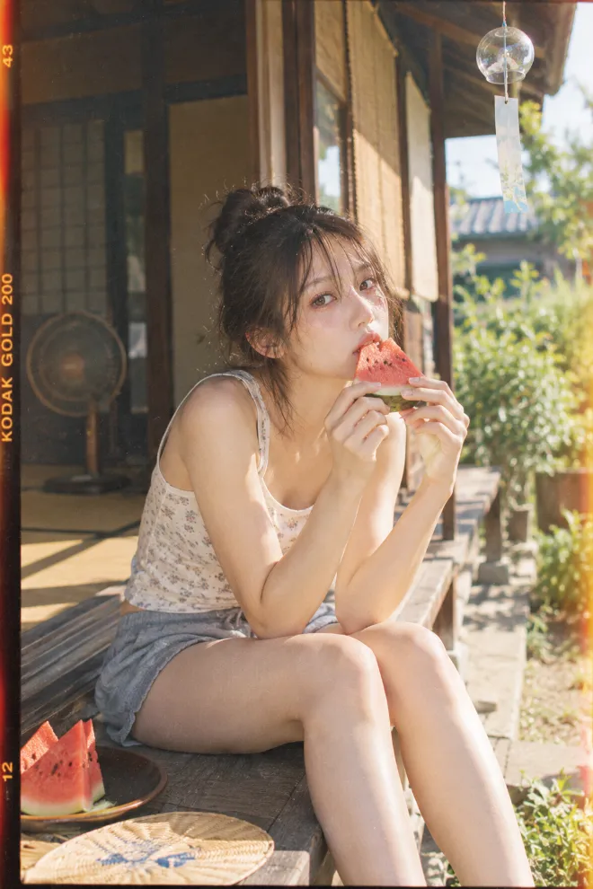 Kodak Gold 200で撮影したようなフィルム写真。ラフな服装の20歳の日本人女性が夏の縁側で風鈴のそばに座り、スイカを食べている。